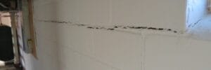 A white painted basement cinder block wall with a visible horizontal crack running through the middle and a small window on the right.