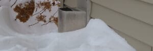 A metal vent partially covered in snow is attached to the side of a beige house. Nearby, some dried plants are also partially buried under the snow.
