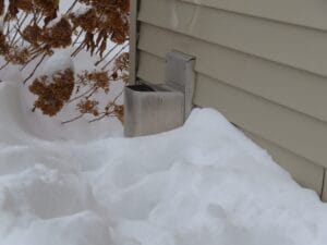 A metal vent partially covered in snow is attached to the side of a beige house. Nearby, some dried plants are also partially buried under the snow.