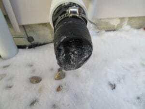 A black pipe covered in ice and snow is seen outdoors near a building wall. The ground around the pipe is also snowy, with some visible rocks.