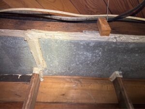 White paper-like tape along the seams of a metal heating duct between wood floor joists in a basement