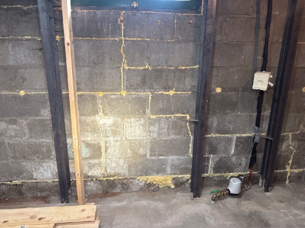 Concrete block basement wall with yellow spray foam pushed into many cracks and mortar joints.