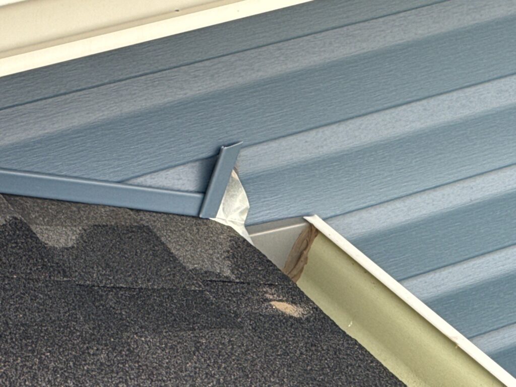 Small metal kick-out flashing at the bottom of a roof-wall joint above a gutter, on blue vinyl siding