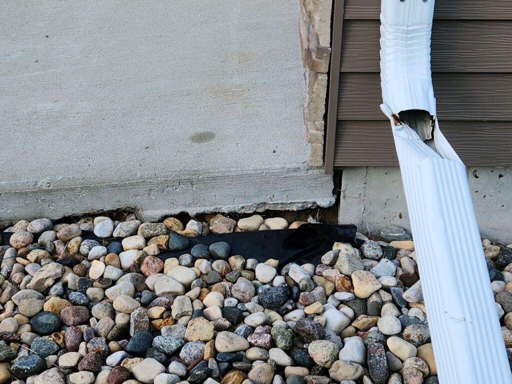 Gap under a concrete driveway slab next to the house foundation and a downspout