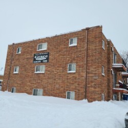 Three-story brick apartment building with snow-covered ground and trees. Visible signage reads "Grace Properties" with contact information and some balconies on the side of the building.