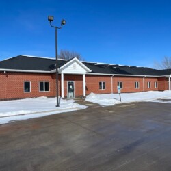 A single-story red brick building with a black roof and white trim is set against a clear blue sky. Snow covers the ground around the building and a parking lot is visible in the foreground.