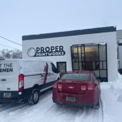 A white van and a red sedan are parked in front of a building labeled "Proper Printworks" on a snowy day. The van has "Trust the Norsemen" and "Nordic Insulation" written on it.