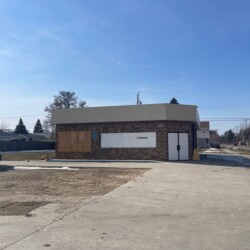 A small, vacant brick building with boarded windows and a blank sign sits on a concrete lot. A service van is parked to the left of the building, and there are leafless trees in the background.