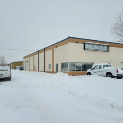 A beige industrial building with number 1721, a snow-covered parking lot, and two parked vehicles in a snowy landscape with bare trees.