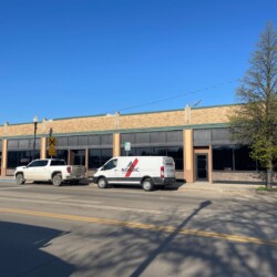 A beige commercial building with several storefronts, including a white van and a white truck parked in front on a sunny day. A tree stands on the right side of the image.