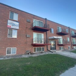 A two-story brick apartment building with balconies. The sign reads "Cottonwood Apartments." The building is surrounded by grass and concrete walkways.