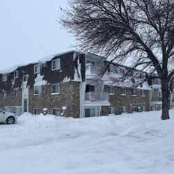 An apartment building with snow-covered roofs and surroundings, a parked white car, and bare trees nearby on a snowy day.