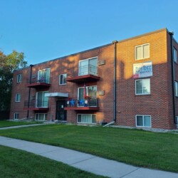 A three-story red brick apartment building with balconies and a "For Rent" sign on the lawn and a "Elm Tree Apartments" sign on the facade.