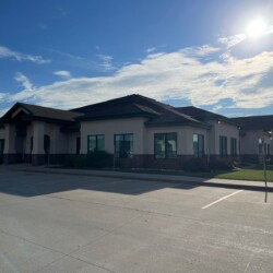 A single-story building with a dark roof, large windows, and a light exterior under a sunny sky, surrounded by an empty paved parking lot.