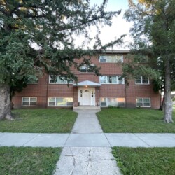 A two-story red brick apartment building with a central entrance, surrounded by two large evergreen trees and a concrete pathway leading to the front door.