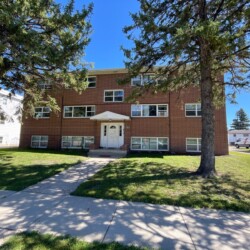 A three-story brick apartment building with white trim, located between two large pine trees, viewed from the sidewalk on a sunny day.
