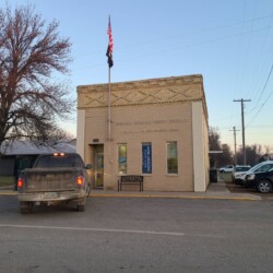 A small brick building with a U.S. flag is the United States Postal Service office. A pickup truck is parked in front. Bare trees and other buildings are visible in the background.