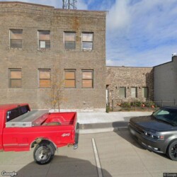 A red pickup truck and a dark SUV parked in front of a multi-story brick building with boarded-up windows and a sidewalk out front.