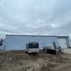 A large industrial warehouse with a corrugated metal exterior, surrounded by trailers and equipment, under an overcast sky.