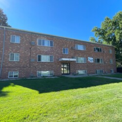 A three-story brick apartment building with multiple windows, a central entrance, and a manicured lawn in the foreground on a sunny day.