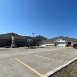 A large, two-story building with an attached garage, parking lot, and several vehicles. The sky is clear and blue.