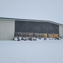 A large building with a dark central section and light-colored sides sits under a snowy sky. The ground is covered with snow, and a set of windows shows light from within.