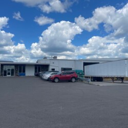 A parking lot with several cars and a large white trailer in front of a warehouse building under a partly cloudy sky.