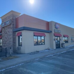 A Dairy Queen Grill & Chill restaurant with beige and red exterior, seen on a sunny day. The building has large windows and outdoor seating. A parking lot is visible in the foreground.