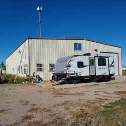 A grey RV is parked outside a beige industrial building with a ladder leaning against the side. A white pickup truck with the text “TRUST THE NORSEMEN” is parked nearby. The sky is clear and blue.
