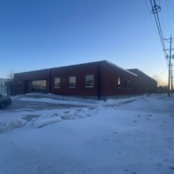 A single-story brick building, surrounded by snow-covered ground, with several windows and a car parked nearby, is shown at dusk. Power lines run along the street.