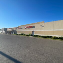 A large beige building with signs for a liquor store and other retailers. The parking lot is empty and the sky is clear and blue.