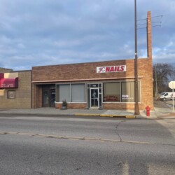 A single-story nail salon with a "JC Nails" sign on the facade, located between a bar and an empty lot. The sidewalk is empty and a fire hydrant is visible near the entrance.