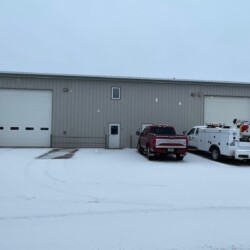 A large gray warehouse with two trucks parked in front on a snowy day. The building has two large garage doors and a smaller entry door in the center. Snow covers the ground and some surrounding trees are visible.