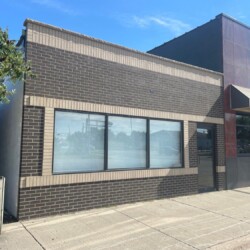 A brick building with large window panels on the front facade, sits adjacent to another building on a sunny day. A tree and trash can are visible on the sidewalk to the left.