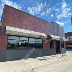 A red and black building with large windows and two awnings stands on a corner street under a blue sky with clouds. A streetlamp and a road sign are visible on the right side.