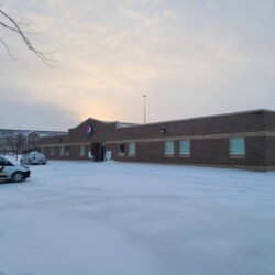 A brick building with a Pepsi logo is surrounded by snow. A white van and a car are parked in the snowy lot. Leafless trees frame the image while the sky appears overcast.