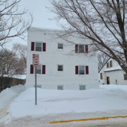 A three-story white building with red shutters is surrounded by snow. Bare trees stand nearby, and a "No Parking Any Time" sign is visible in the foreground.