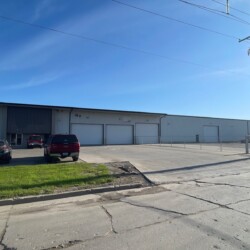 Large industrial warehouse with a metal facade, several large garage doors, and a fenced-in area. A few vehicles are parked in front on a paved area. Overhead utility lines are visible against a clear sky.