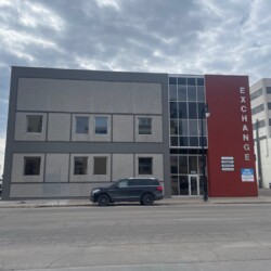 A two-story building with a gray facade and a red section displaying the word “EXCHANGE”. A black SUV is parked in front. The sky is overcast. The street and nearby buildings are visible.