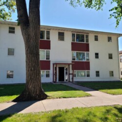 A three-story white apartment building with red accents, surrounded by grass and trees, with a sidewalk leading to the entrance.