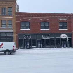 A snowy street with a white van parked in front of a building with a "623" sign, various businesses, and snow-covered ground.