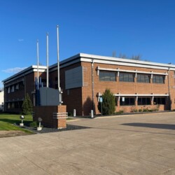 A two-story brick building with three flagpoles in front and a parking lot on one side, set against a clear blue sky and greenery.