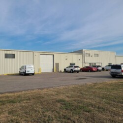 A light beige industrial building with multiple parked vehicles in front, including a white van, a red car, and several white SUVs. The sky is clear and blue.