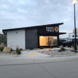 A modern yoga studio named "Haute Yogis" with large windows, located on a corner with surrounding rocks and plants. An SUV is parked near the studio.