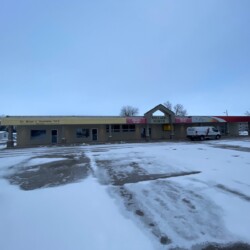 A small commercial building with a dentist's office, a nail salon, and a U.S. Postal Service. The ground is covered with patches of snow. A white van is parked on the right side.