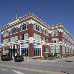 A three-story red brick building with many windows and a "MercyOne Health & Fitness Center" sign. Two black cars are parked in front, and the building is situated on a corner lot with a clear sky above.