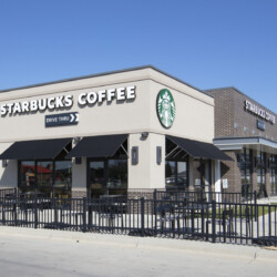 A standalone Starbucks Coffee building with a drive-thru and outdoor seating area, situated in a commercial area under a clear blue sky.